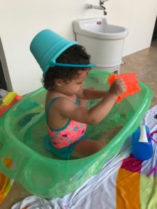 A baby wearing a bathing suit  and a blue sand bucket on her head plays in a bathtub. 