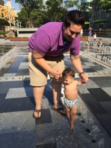 A woman helps a baby walk in a water fountain.